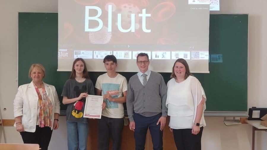 Five people stand in front of a green board with a projector screen displaying the word 'Blut'. A man holds a certificate, while the others smile.