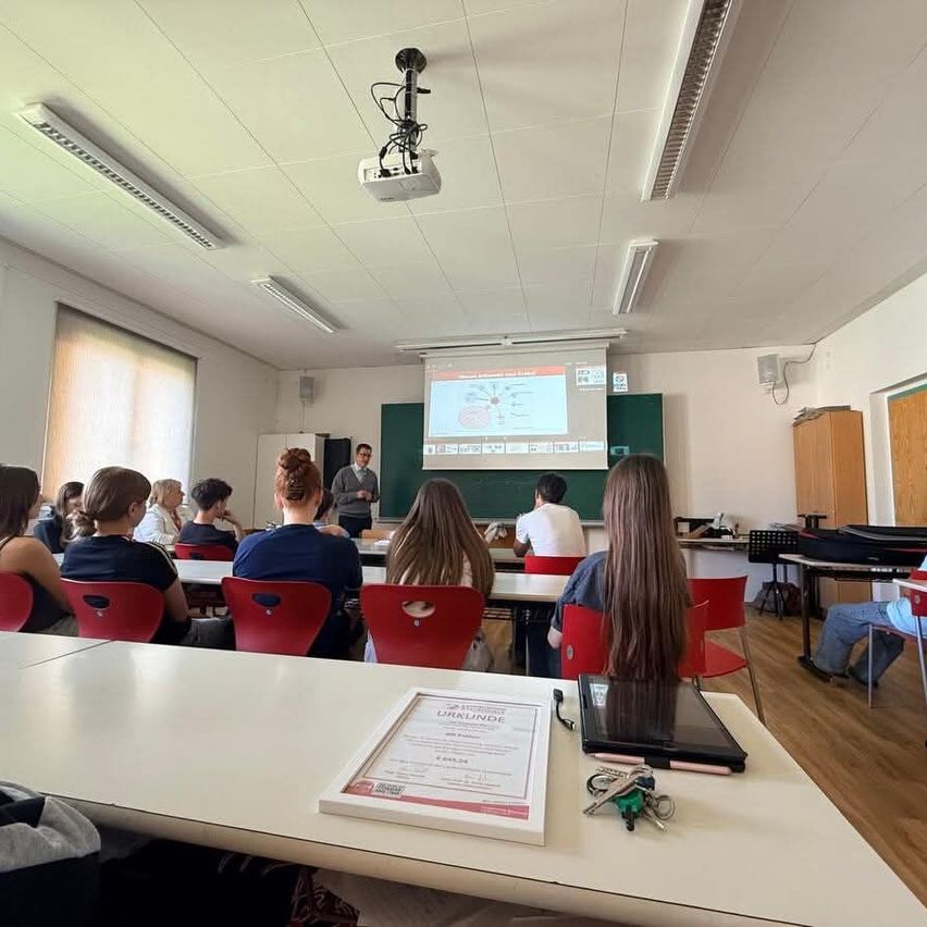 A classroom with students and a teacher. The teacher is standing and speaking in front of a projector screen displaying a diagram. The room is equipped with a projector and overhead lights.