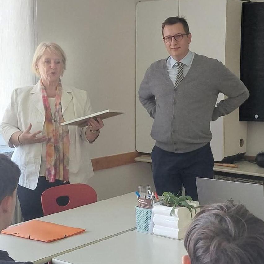 Two people, a man and a woman, are standing in a classroom. The woman is holding a folder while speaking. A boy is sitting in front of them, and a table with books and a laptop is nearby.