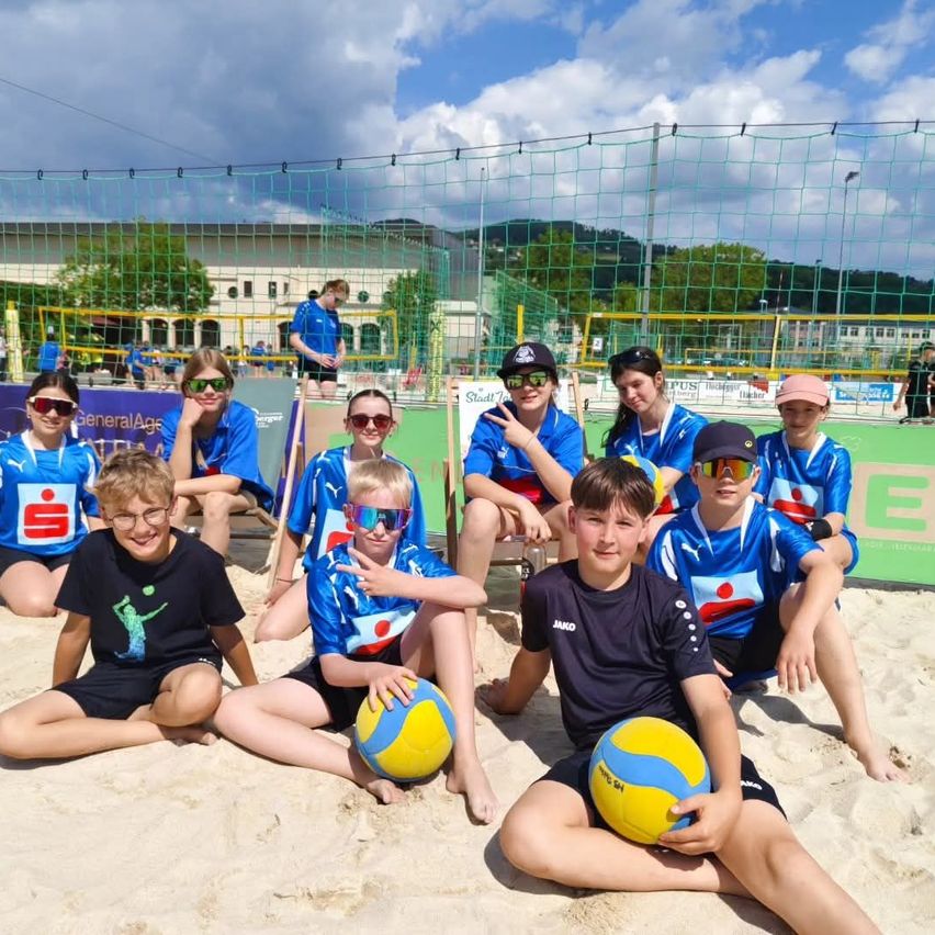 Eine Gruppe von Kindern in passenden blauen Trikots sitzt auf einem sandigen Strand und hält Beachvolleyballbälle. Sie tragen Sonnenbrillen und scheinen für ein Foto zu posieren.