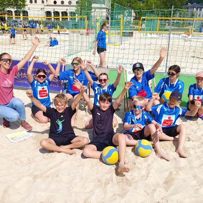 Eine Gruppe junger Athleten posiert für ein Foto am Strand mit einem Volleyballnetz im Hintergrund. Sie tragen blaue Trikots und halten Bälle.