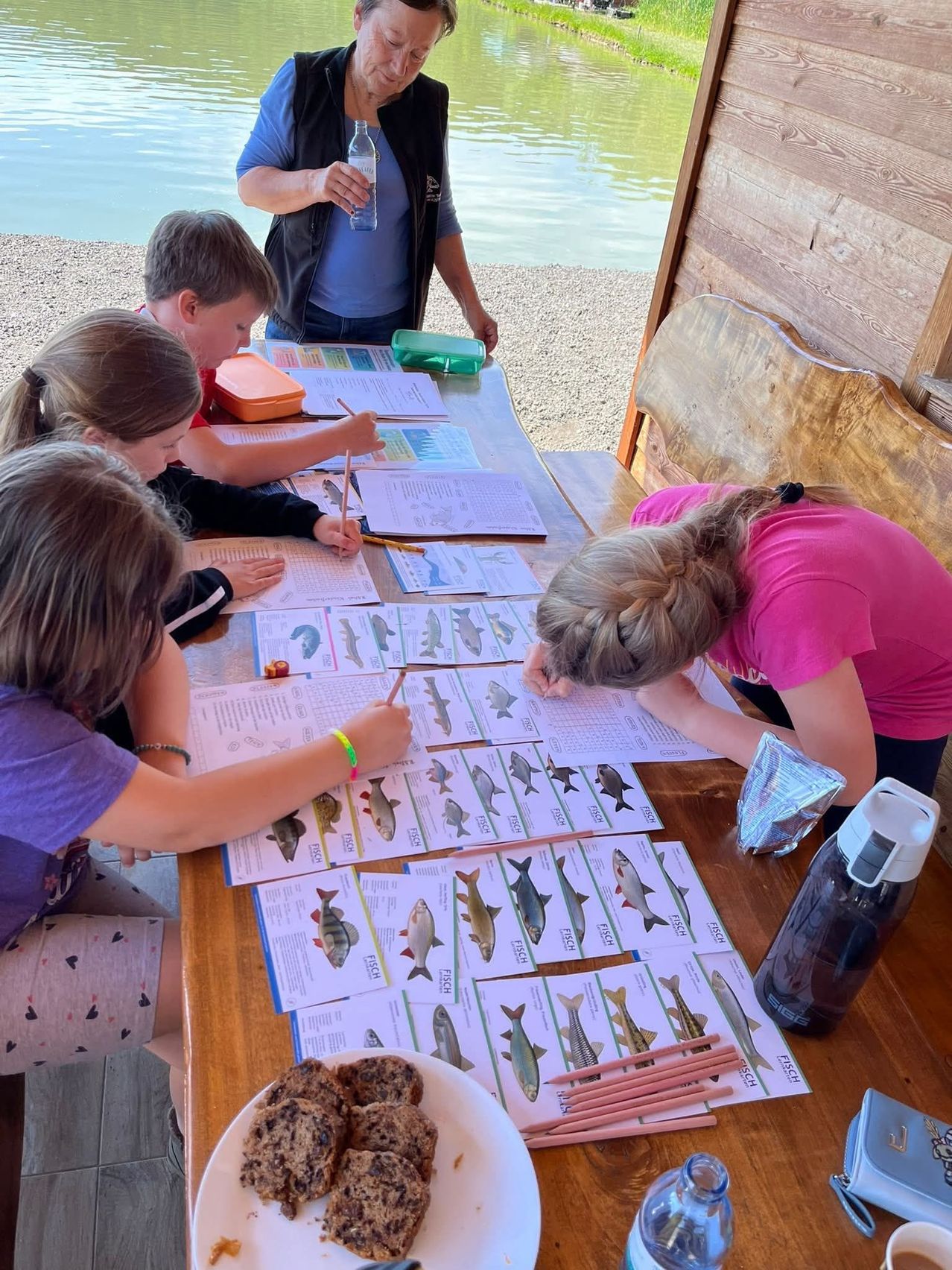 Kinder studieren Fische an einem Tisch am See. Eine Person im Hintergrund hält eine Wasserflasche. Sie haben Fischkarten, Papiere und Stifte auf dem Tisch.