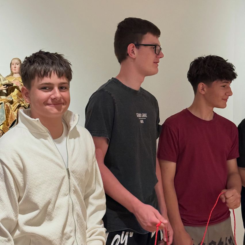 Drei junge Männer stehen zusammen in einem Raum, lächeln und schauen in die Kamera. Der Mann in Weiß trägt einen hellen Mantel. Hinter ihnen steht eine goldene Statue einer Frau.