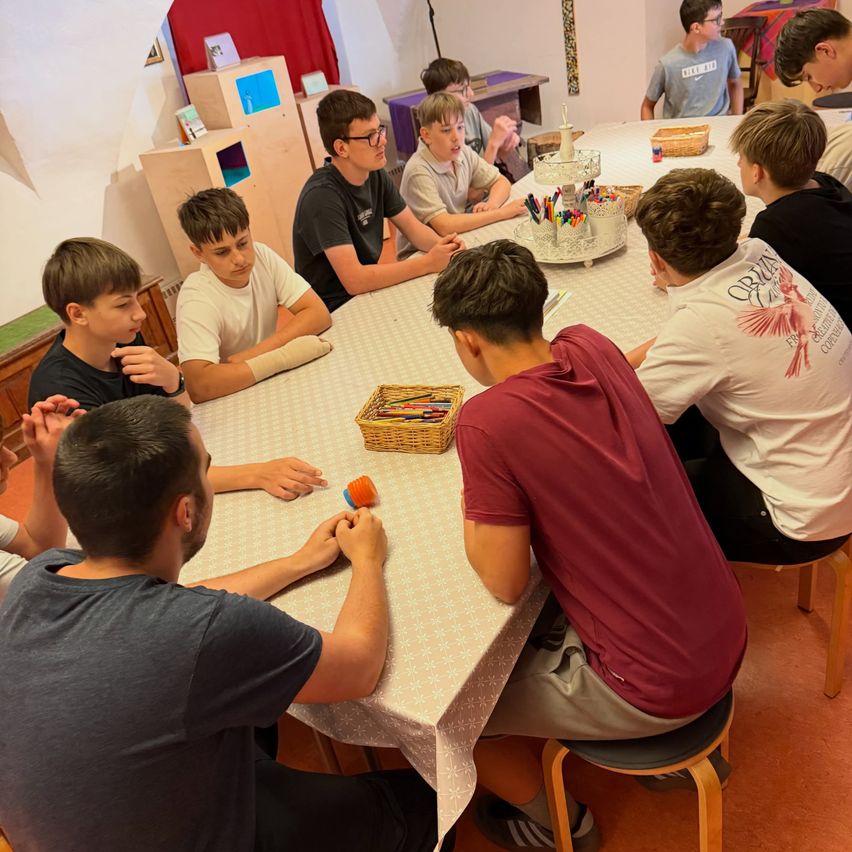 Mehrere Jungen und ein Mann sitzen um einen Tisch und beschäftigen sich mit einer Aktivität. Der Tisch ist mit einem Tuch bedeckt und darauf befinden sich verschiedene Gegenstände, darunter ein Korb mit farbigen Bleistiften.