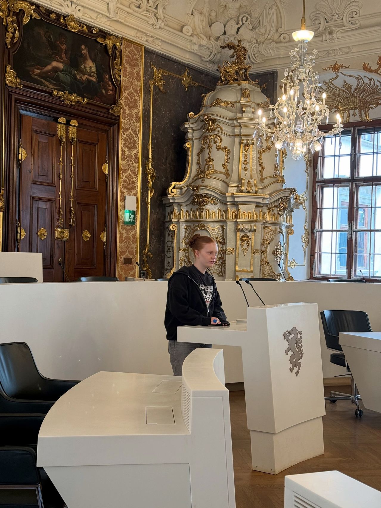 Eine junge Frau steht an einem Rednerpult in einem großen Saal mit prächtigem Goldrand, einer Kronleuchter und einem Gemälde. Sie steht vor einem Mikrofon mit einem Löwenlogo auf dem Pult. Hinter ihr ist ein großer, verzierter Kamin sichtbar.