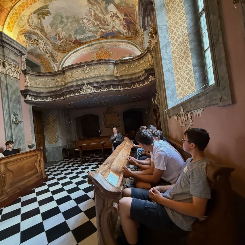 Eine Gruppe von Menschen sitzt auf hölzernen Bänken in einer Kirche und hört einer Frau zu. Der Raum hat einen schwarz-weiß karierten Boden, Holzbänke und kunstvolle Dekorationen.