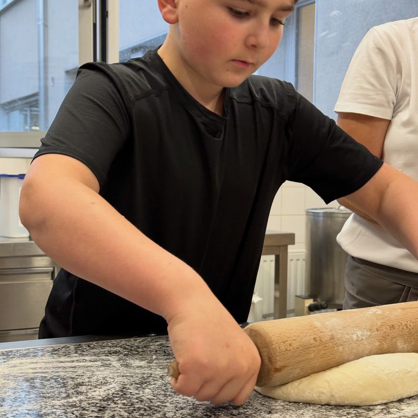 Ein junger Junge rollt Teig auf einem Arbeitstisch mit einem Holzwalzen. Eine andere Person steht neben ihm und schaut zu.
