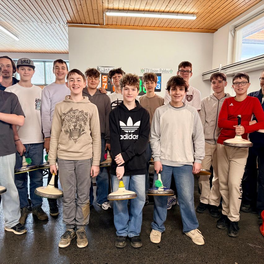 Eine Gruppe junger Männer und Jungen posiert für ein Foto, während sie Curlingbesen halten. Sie befinden sich in einem Raum mit einer hölzernen Decke und weißen Wänden. Einige tragen Turnschuhe, andere Stiefel.