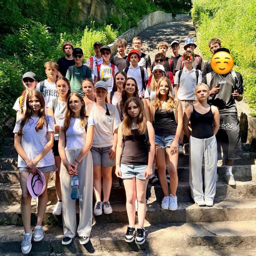 Eine Gruppe von Schülern in Freizeitkleidung, einige tragen Hüte und Brillen, stehen auf Stufen. Ein Schüler hält einen Hut und eine Flasche. Ein lächelndes Emoji ist überlagert.
