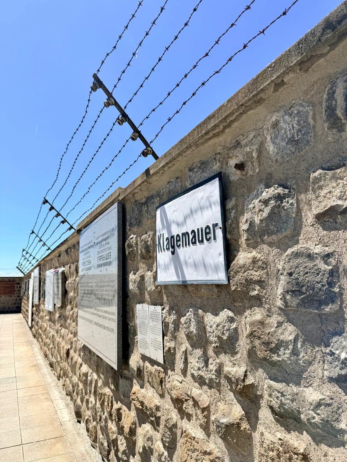 Eine Steinmauer mit Stacheldraht oben, ein Schild mit 'Klagemauer' und weitere Plaketten befestigt. Die Mauer ist unter einem klaren blauen Himmel.