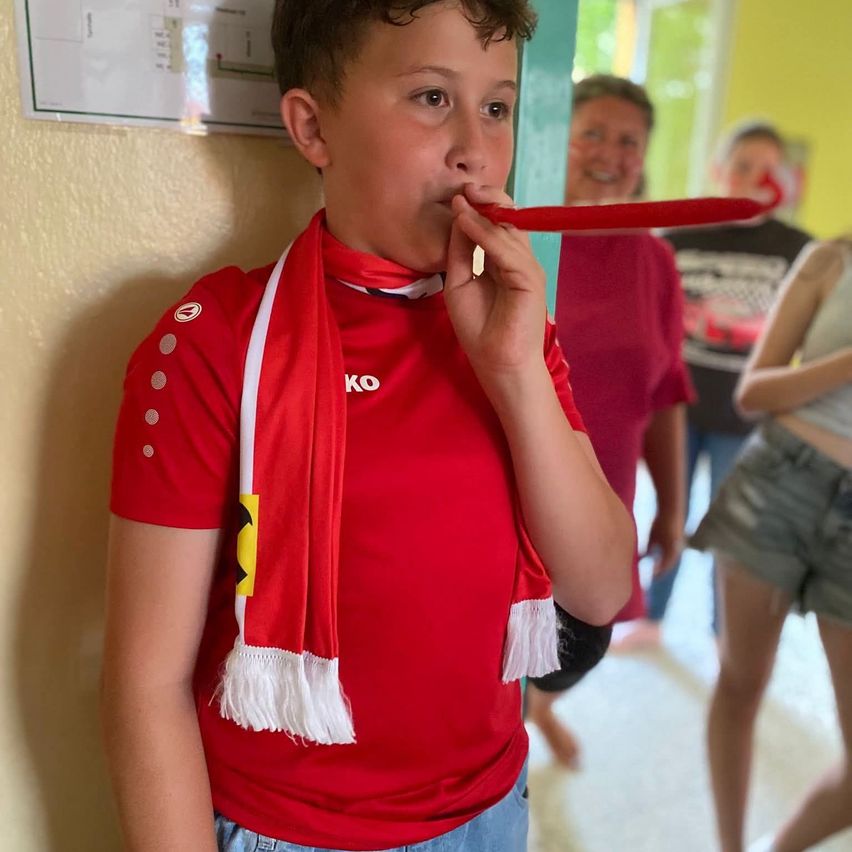 Ein junger Junge in einem roten Trikot und Schal bläst in einem Flur in eine rote Hupe. Dahinter lächelt eine Frau und eine weitere Person schaut zu. Eine Anschlagtafel ist an der Wand angebracht.