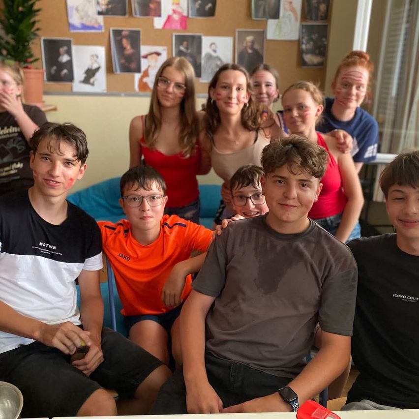 Eine Gruppe junger Personen in einem Klassenzimmer, einige mit Brille, einer mit Halskette, und einige mit Gesichtsbemalung, posieren für ein Foto.