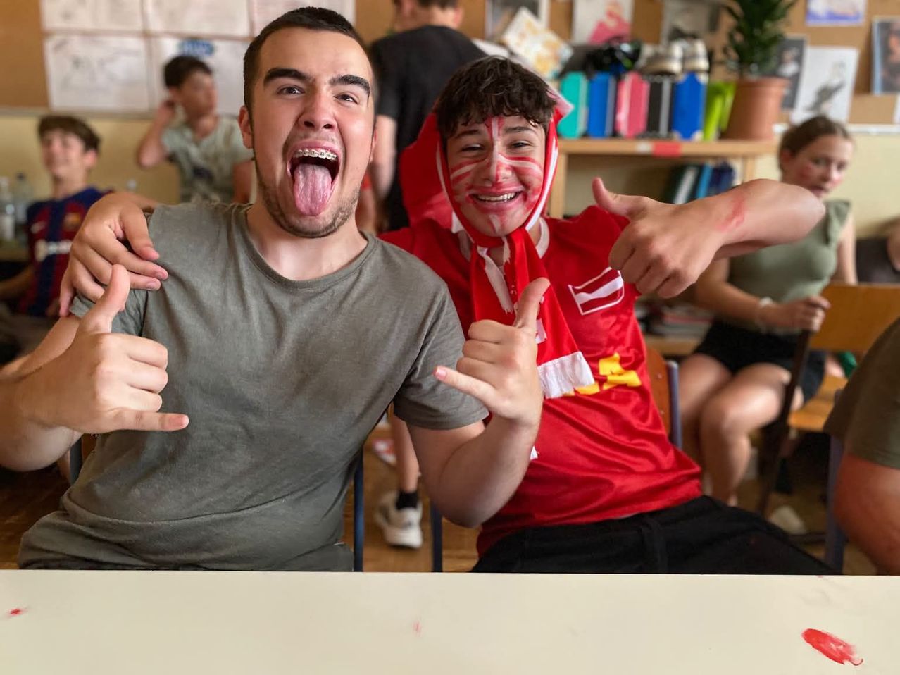 Zwei junge Männer sitzen zusammen, einer mit Zahnspange, der andere mit rotem Farbanstrich im Gesicht, beide machen lustige Grimassen und Gesten in einem Klassenzimmer.