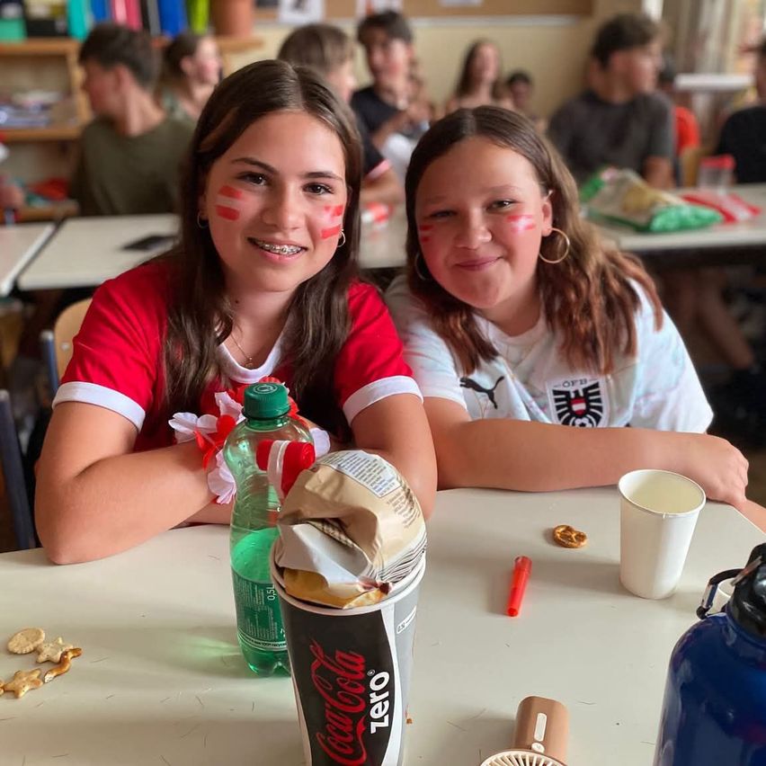 Zwei Mädchen sitzen in einem Klassenzimmer an einem Tisch, lächeln mit rotem Gesichtsschminke. Eine trägt ein rotes Shirt, die andere ein weißes Shirt mit Logo. Ein Coca-Cola-Becher, eine Flasche, ein Becher und Snacks sind auf dem Tisch.