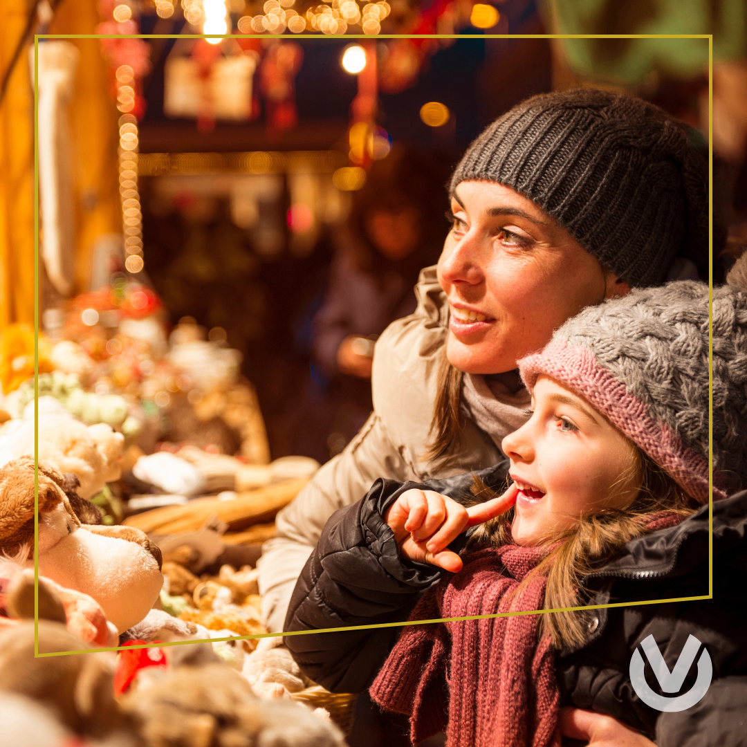 Eine Frau und ein junges Mädchen auf einem Weihnachtsmarkt, sie lächeln und schauen sich Plüschtiere in einem Geschäft an.