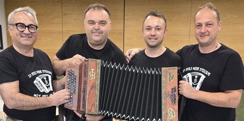 Vier Männer in schwarzen T-Shirts posieren für ein Foto, einer hält ein Akkordeon. Der Mann links trägt eine Brille und der Mann rechts eine Kette.