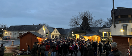 Eine große Menschenmenge versammelt sich bei Dämmerung in einem Freiluftbereich, wahrscheinlich für eine festliche Veranstaltung. Einige tragen Mützen und Winterkleidung, und es gibt kleine Hütten mit Lichtern. Das Gebiet ist von Häusern und Bäumen umgeben, mit einem bewölkten Himmel darüber.