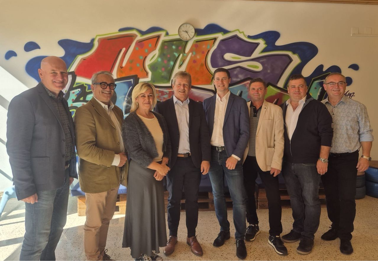 Eine Gruppe von sechs Personen steht vor einer mit Graffiti bedeckten Wand. Sie sind alle formell gekleidet, möglicherweise für eine geschäftliche Veranstaltung. Die Wand hinter ihnen ist farbenfroh und hat Graffiti darauf.