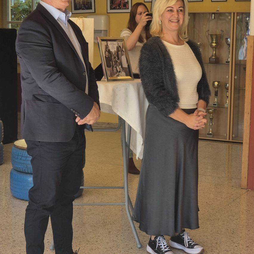 Ein Mann und eine Frau stehen lächelnd nebeneinander, mit einem Fotorahmen auf einem Tisch und einer Frau, die ein Foto macht im Hintergrund. Dahinter steht ein Regal mit Trophäen.