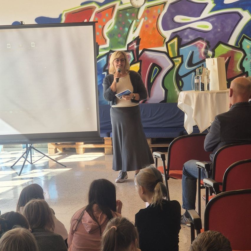 Eine Frau steht vor einer Tafel und spricht in ein Mikrofon. Sie hält ein Buch. Dahinter eine Wand mit Graffiti-Kunst. Ein Tisch mit Flaschen steht in der Nähe. Ein Mann und mehrere Kinder sitzen auf Stühlen.