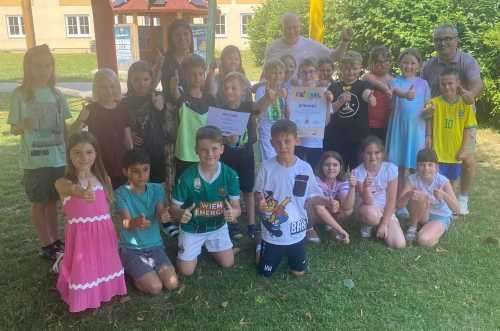 Eine Gruppe von Kindern und Erwachsenen posiert für ein Foto auf einer Rasenfläche mit einem Gebäude und Bäumen im Hintergrund. Einige halten Urkunden, und alle zeigen Daumen hoch.