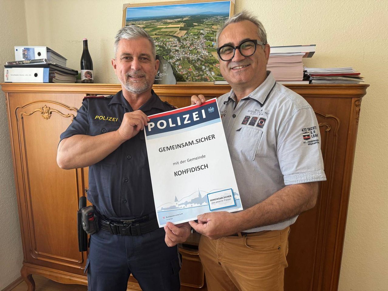 Zwei Männer stehen drinnen, einer in Uniform, und halten ein großes weißes Schild mit der Aufschrift 'POLIZEI GEMEINSAMSUCHER mit der Gemeinde KOHFIDISCH'. Sie lächeln für die Kamera, mit einem Holzschrank und einem gerahmten Bild dahinter.