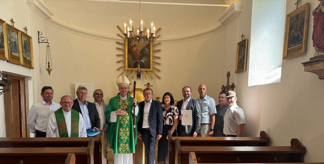 Eine Gruppe von Menschen steht in einer Kirche. Ein Priester hält einen Stab und trägt eine grün-weiße Robe. Eine Frau hält ein weißes Papier. Zwei Männer tragen Hüte.