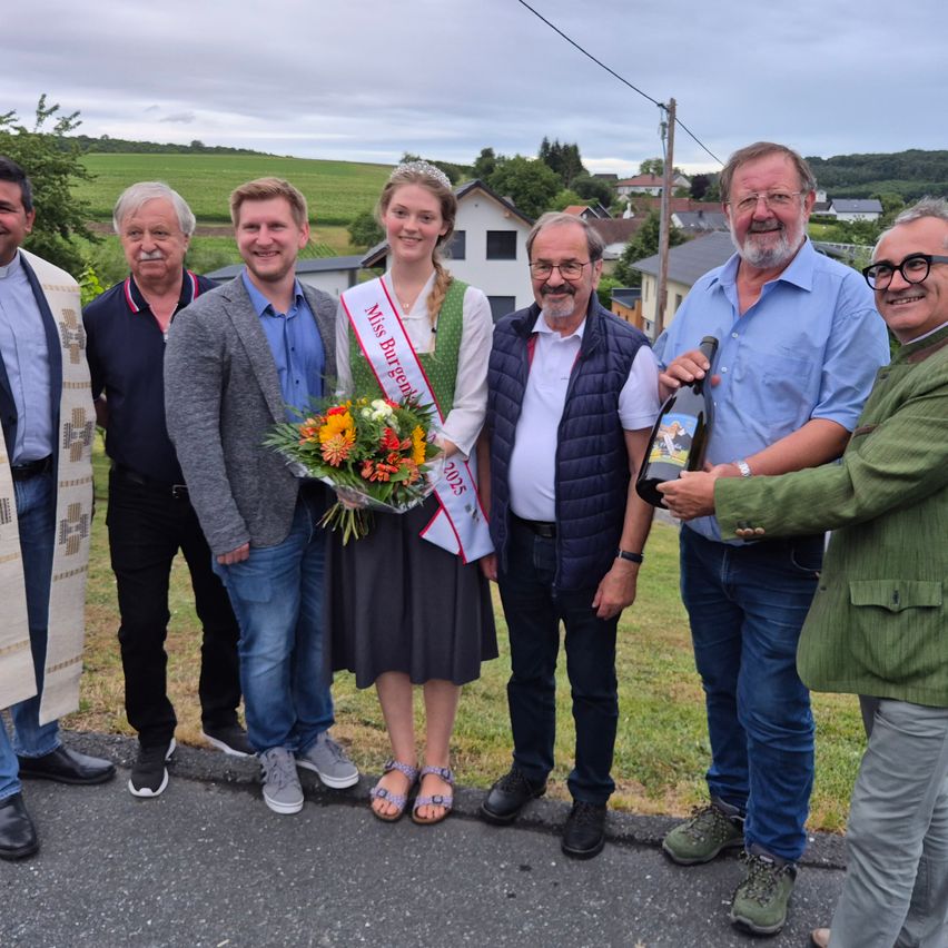 Eine Gruppe von Menschen posiert für ein Foto im Freien. Eine Frau mit einem Band hält einen Blumenstrauß. Ein Mann hält eine Flasche Wein. Hinter ihnen ist ein grasbewachsenes Feld mit Häusern und Bäumen.