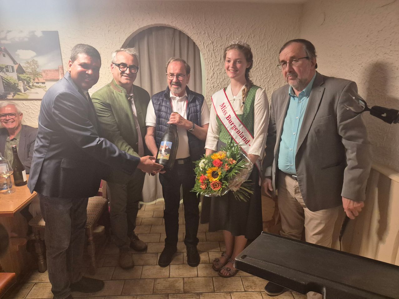 Fünf Männer und eine Frau, gekrönt als Miss Burgenland, stehen drinnen. Der Mann auf der linken Seite hält eine Flasche und einen Blumenstrauß. Der Mann in der Mitte hält eine Flasche. Die Frau hält einen Blumenstrauß.
