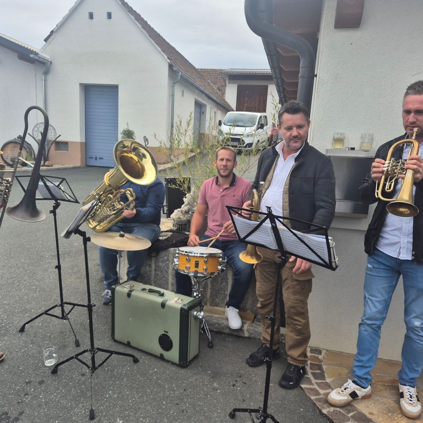 Vier Männer stehen auf einem Parkplatz und spielen verschiedene Musikinstrumente. Ein Mann hält eine Trompete, ein anderer spielt Schlagzeug, und die anderen spielen Tuba und Posaune.