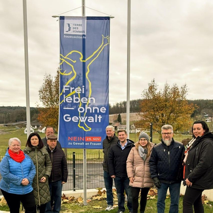 Gruppenfoto mit einem Banner, das 'Frei leben ohne Gewalt' sagt. Die Leute tragen Winterkleidung und stehen auf Gras. Bäume und Häuser sind im Hintergrund.