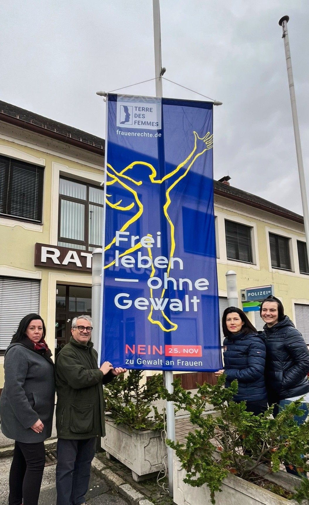 Vier Personen stehen vor einem blauen Banner, das 'Frei leben ohne Gewalt' mit einem Logo oben, einem Datum und einem Slogan 'Nein zu Gewalt an Frauen' zeigt. Das Gebäude hinter ihnen hat ein Schild 'RAT' und ein Polizeischild.