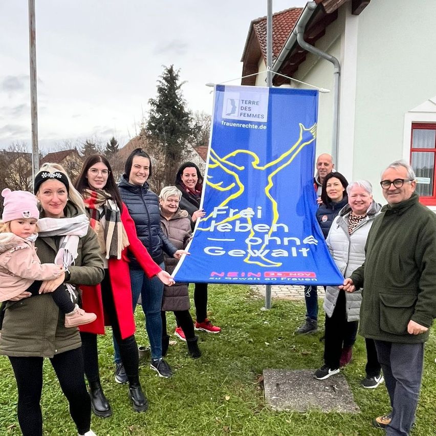 Eine Gruppe von Menschen steht vor einem Haus mit einer blauen Fahne, die eine Silhouette einer Frau zeigt. Sie lächeln und posieren für ein Foto.