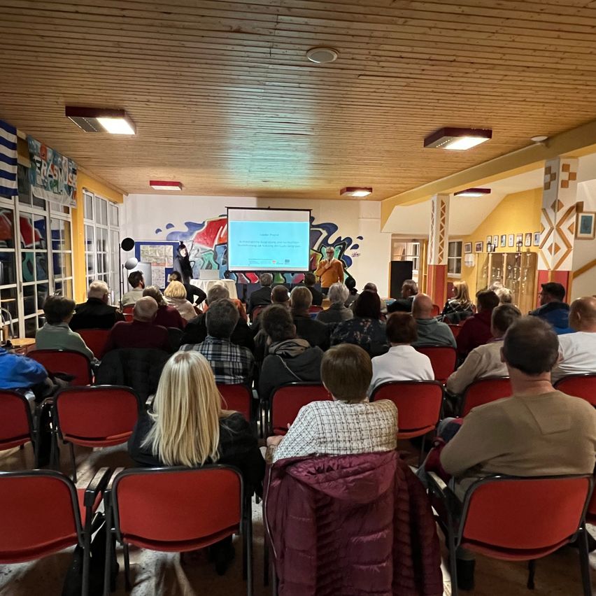 Ein Konferenzraum mit Holzdecke und gelben Wänden. Eine Person steht an einem Rednerpult, während eine andere an einem Schreibtisch sitzt. Eine Projektionsleinwand zeigt eine Präsentation. Die Teilnehmer sitzen in roten Stühlen.