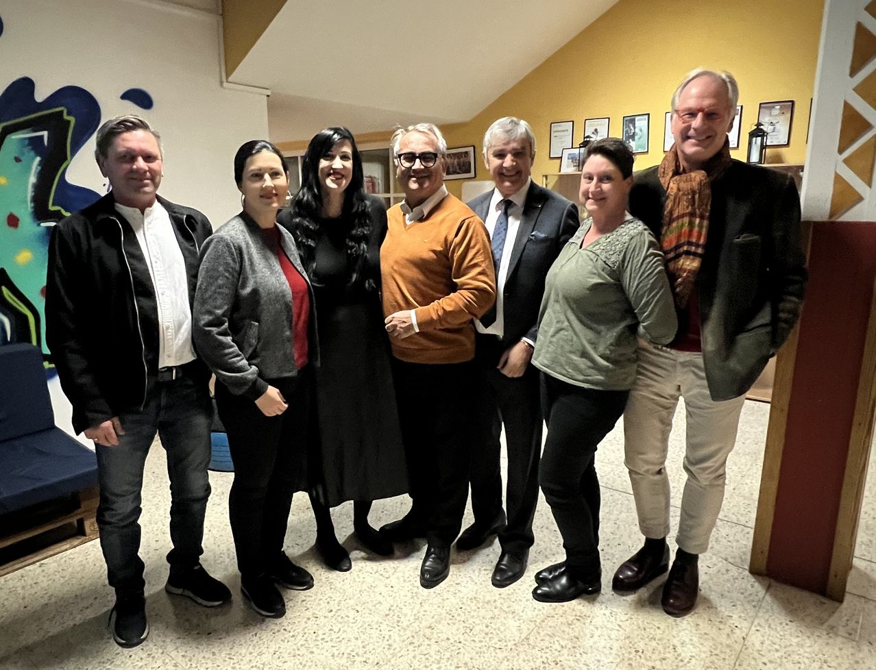 Eine Gruppe von sechs Personen steht in einem Raum mit gelben Wänden. Sie lächeln und scheinen für ein Foto zu posieren. Der Mann in der Mitte trägt eine Brille und einen orangefarbenen Pullover.