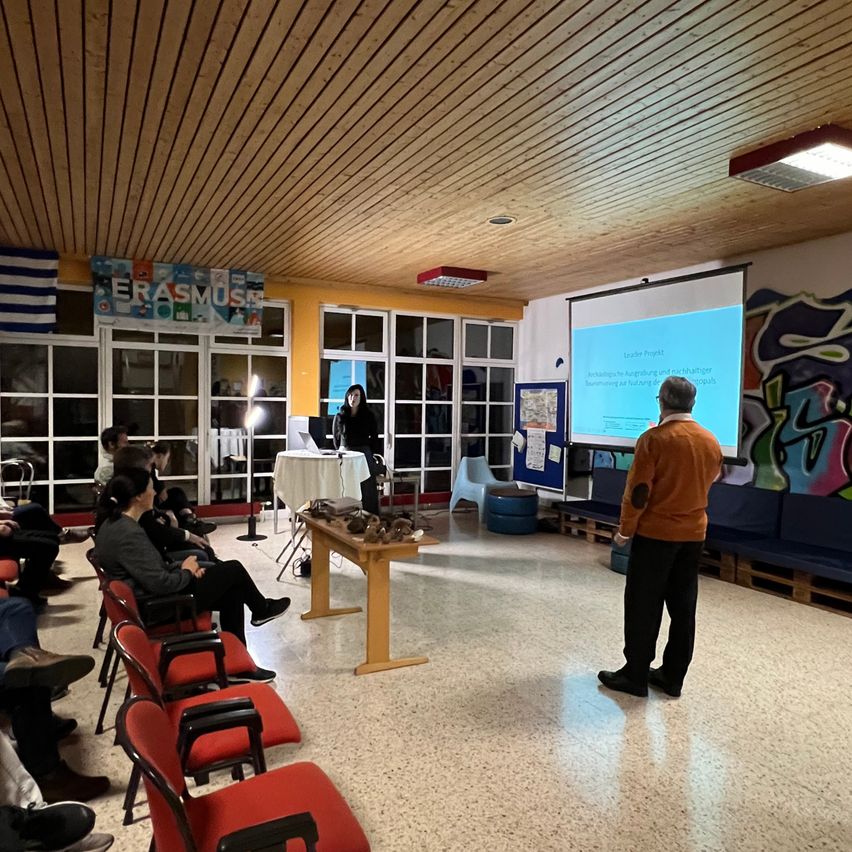 Eine Innenpräsentation mit einer Projektionsleinwand, einem stehenden Mann und einem sitzenden Publikum in roten Stühlen. Eine Frau sitzt an einem Tisch mit Laptop und Mikrofon.