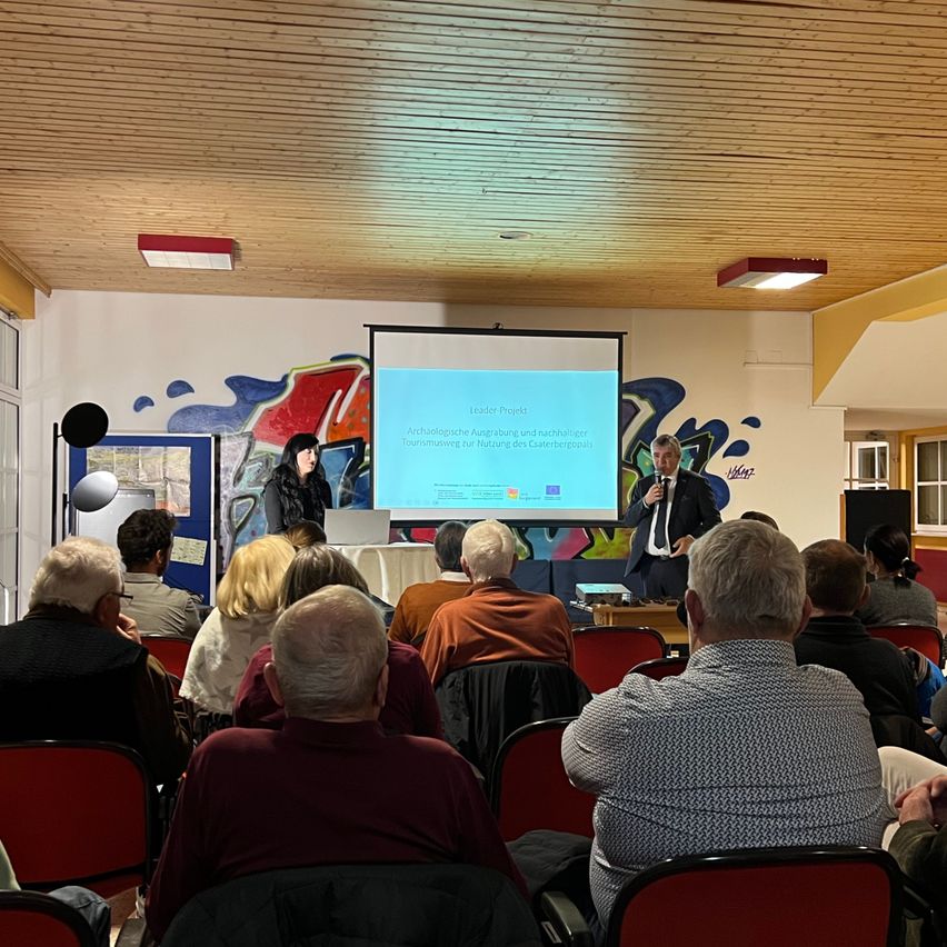 Ein Mann präsentiert eine Folie einem Publikum in einem Raum. Das Publikum sitzt in Reihen, und zwei Personen stehen auf der Bühne. Eine von ihnen hält einen Laptop.