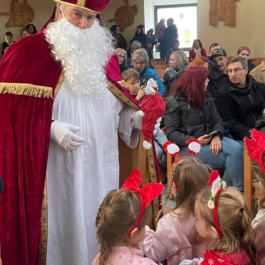 Ein in einem roten Gewand und weißem Umhang gekleideter Weihnachtsmann spricht in einer Kirche mit einer Gruppe von Kindern mit roten Schleifen, während mehrere Erwachsene von hinten zusehen.
