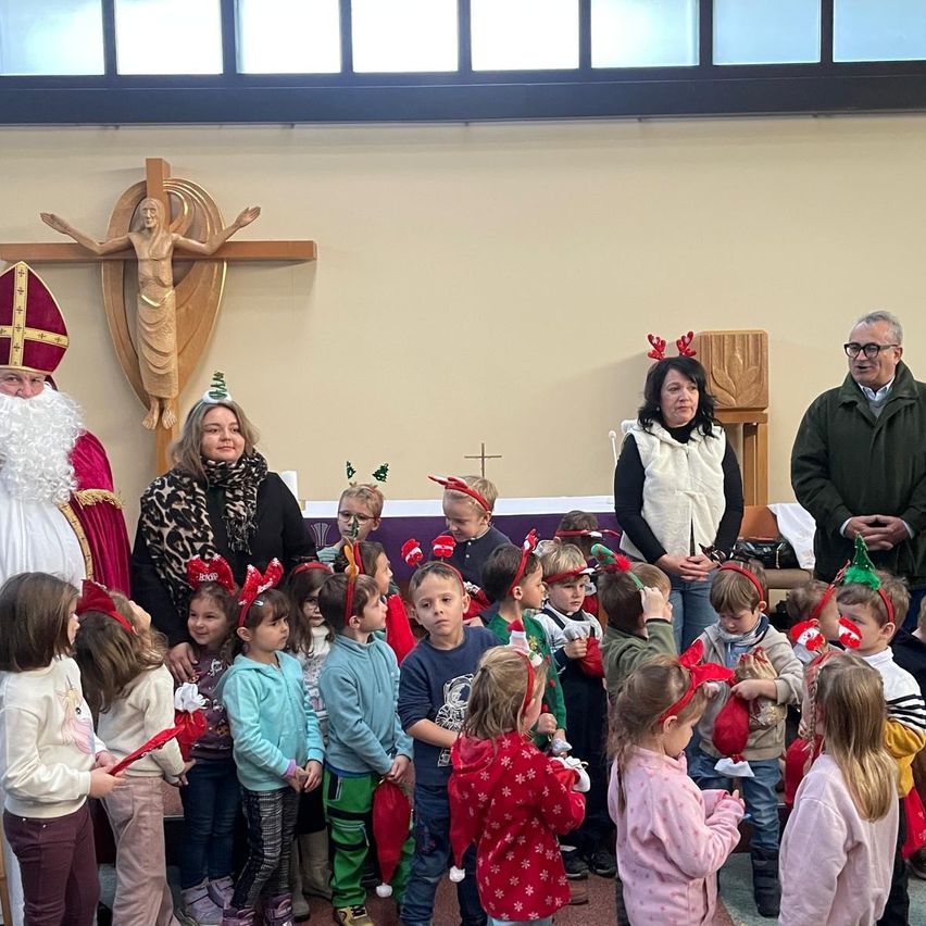Eine Gruppe von Kindern und Erwachsenen versammelt sich in einer Kirche, einige mit Weihnachtsmützen gekleidet, während ein Mann in einer Santa-Claus-Kostüm und eine Frau in einem Leopardenmuster-Schal in der Nähe stehen.
