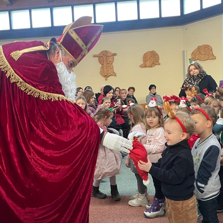 Ein Mann, der als St. Nikolaus verkleidet ist, gibt einem Kind in einer Kirche, umgeben von anderen Kindern und Erwachsenen, ein Geschenk.