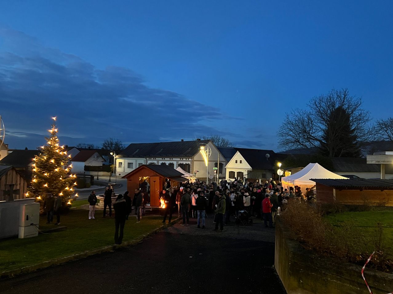 Bei Dämmerung versammelt sich eine Menschenmenge auf einem Stadtplatz, erhellt von festlichen Lichtern und einem Weihnachtsbaum, mit mehreren Zelten in der Nähe.