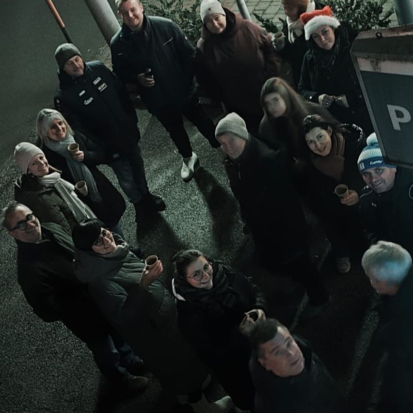 Eine Gruppe von Menschen in Winterkleidung steht im Kreis, einige tragen Mützen, lächeln und halten Tassen. Eine Person trägt einen Weihnachtsmannhut. Die Umgebung scheint im Freien zu sein.