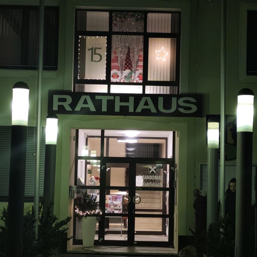 Der Eingang zu einem Gebäude namens Rathaus bei Nacht. Ein Stern ist im Fenster beleuchtet und ein Topf mit Pflanzen steht vor der Glastür.