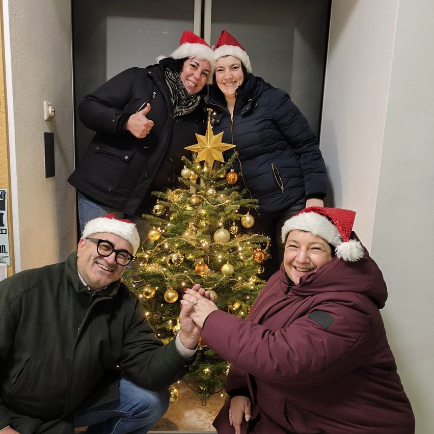 Drei Personen in Winterkleidung stehen vor einem geschmückten Weihnachtsbaum und posieren für ein Foto. Die Frau links gibt einen Daumen hoch, während die Frau rechts die Hand des Mannes vor ihr hält. Der Mann kniet und trägt eine Brille.
