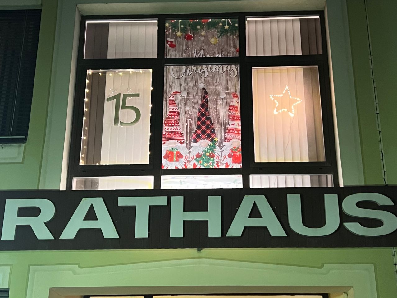 Ein Fenster mit Weihnachtsdekorationen und der Nummer 15, mit Zwergen und einem Stern. Darunter steht ein Schild mit der Aufschrift 'RATHAUS'.