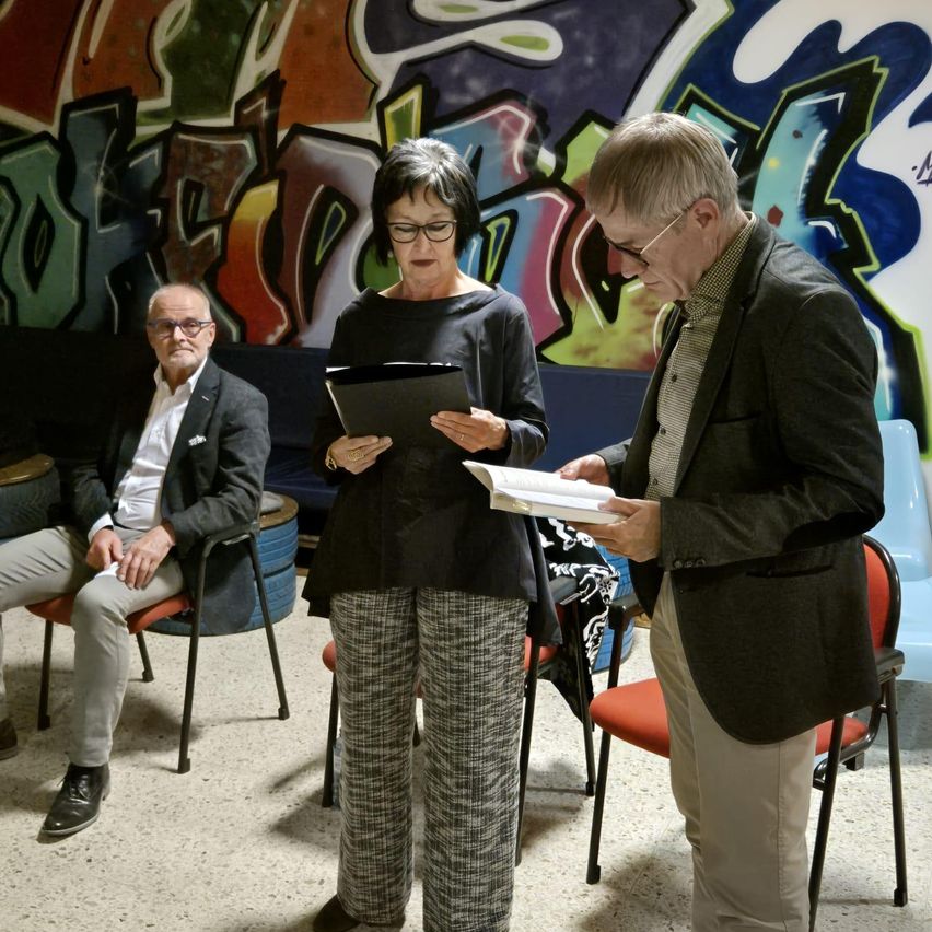 Eine Frau steht zwischen zwei Männern, die ein Buch lesen. Hinter ihr sitzt ein Mann auf einem Stuhl. Die Wände sind mit Graffiti bedeckt.