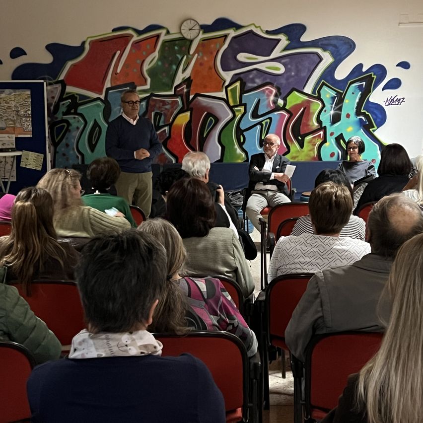 Eine Gruppe von Menschen sitzt in roten Stühlen in einem Raum. Zwei Männer sitzen auf der Bühne, einer steht. Eine Uhr und bunte Graffiti sind an der Wand.