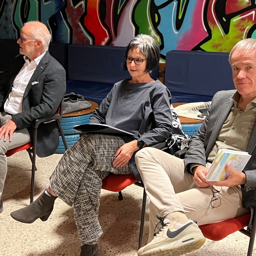 Drei Menschen sitzen in Stühlen, zwei Männer und eine Frau, alle mit Brille. Sie sind in einem Raum mit farbenfrohen Graffiti an der Wand. Die Frau hält eine Mappe, der Mann neben ihr ein Buch.