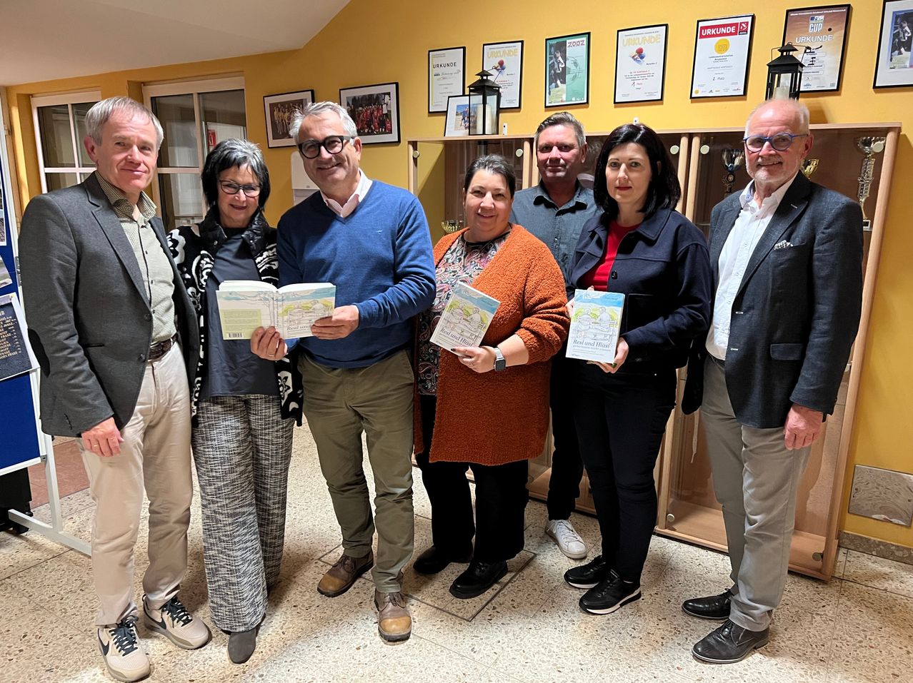 Sieben Personen stehen in einem Raum mit gelben Wänden und Holzregalen, halten Bücher und lächeln für die Kamera. Gerahmte Zertifikate und eine Laterne hängen an der Wand.