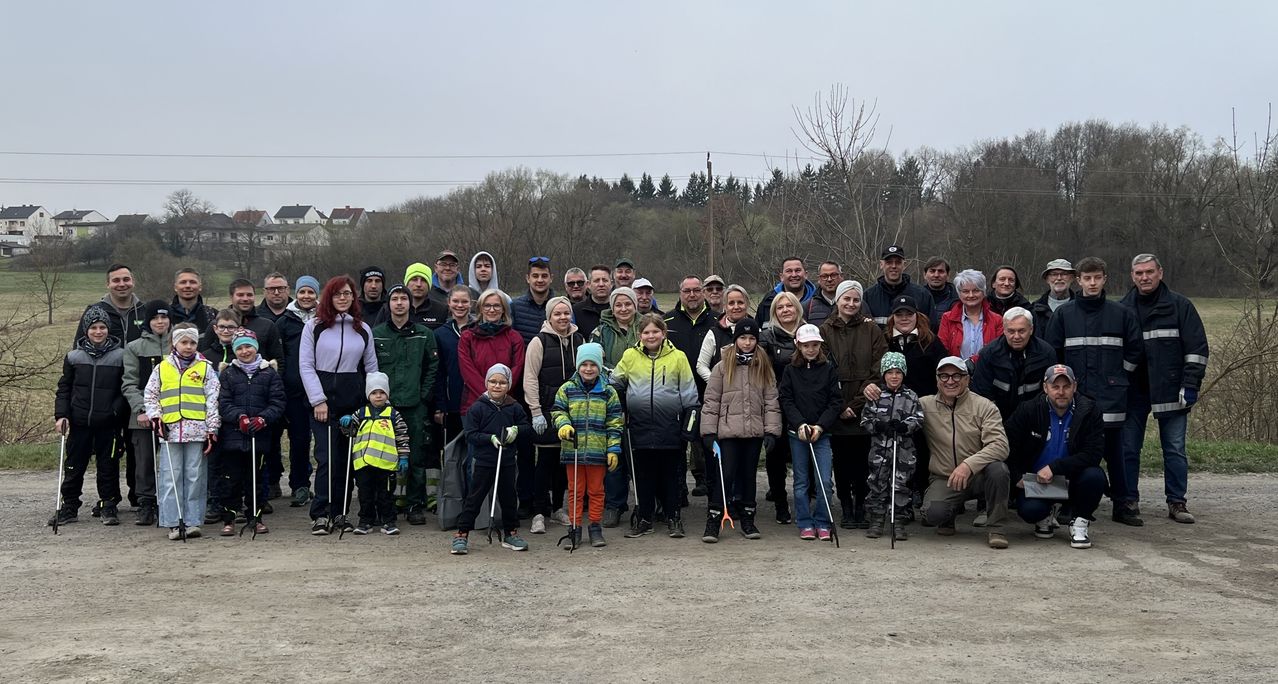 A large group of people stand in a line outside, wearing winter clothing, smiling, and holding sticks. The background is a wooded area with a clear sky.
